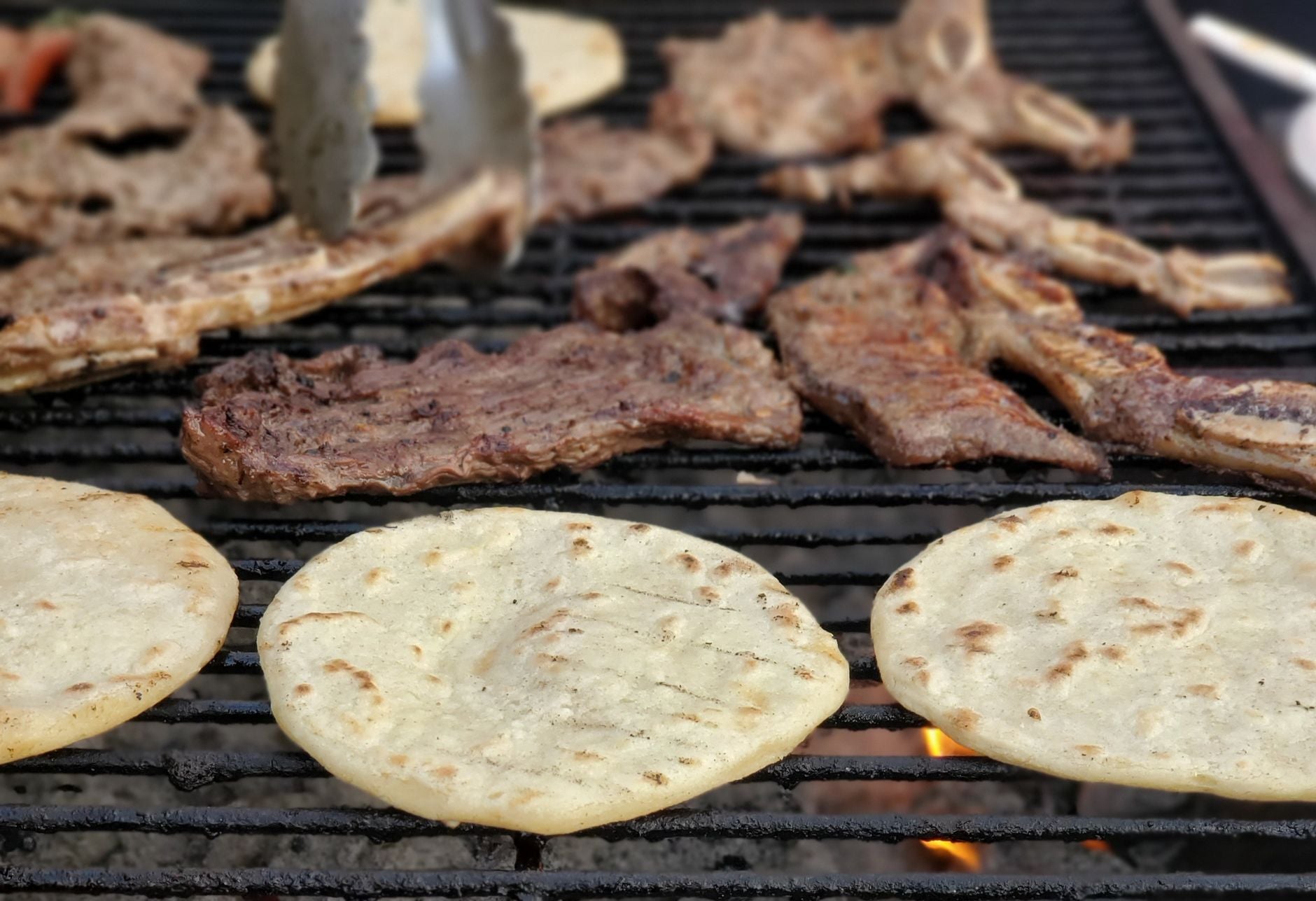 Carne asada and tortillas over charcoal, tongs mid-turn