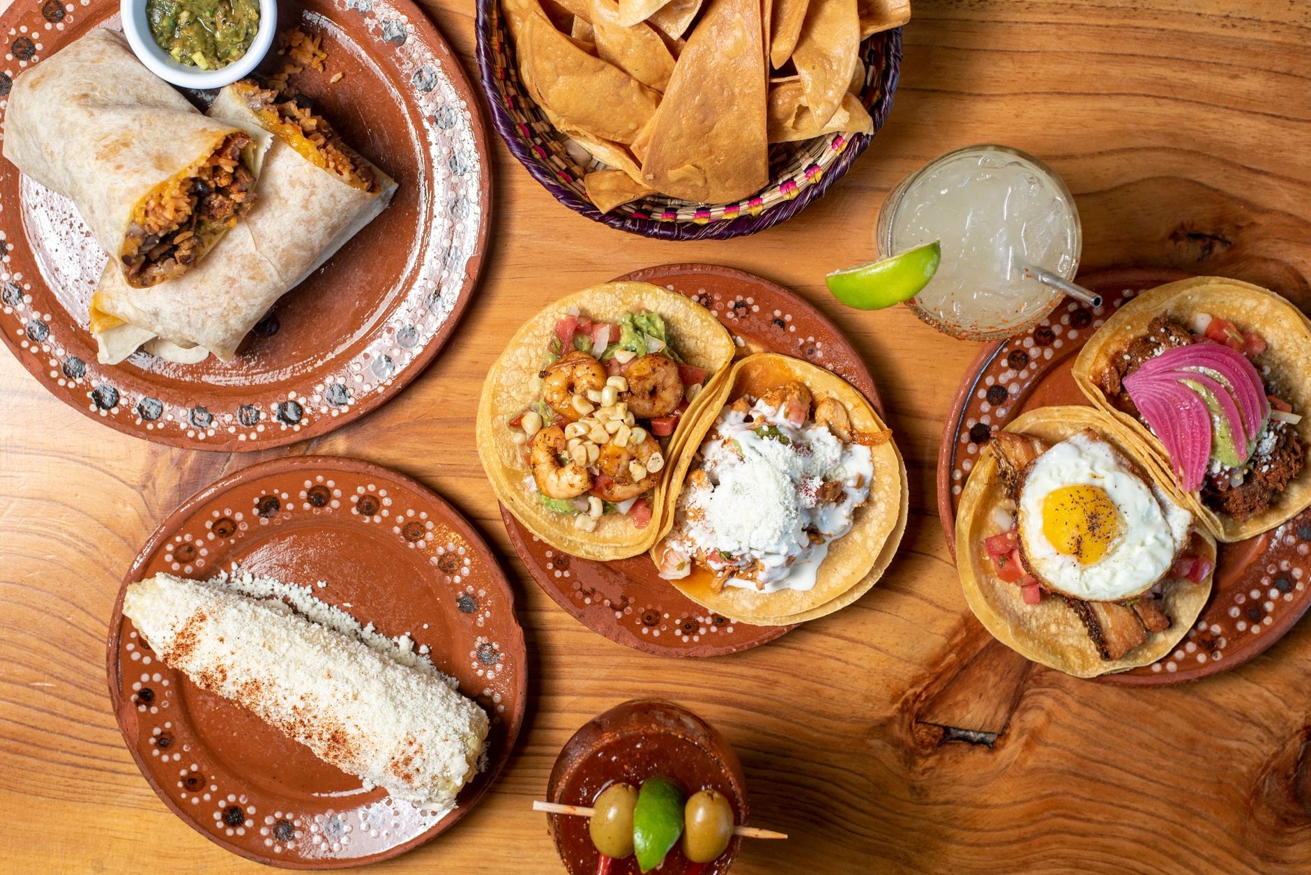 A party spread on clay plates: burritos, street tacos, elote, chips and aguas frescas