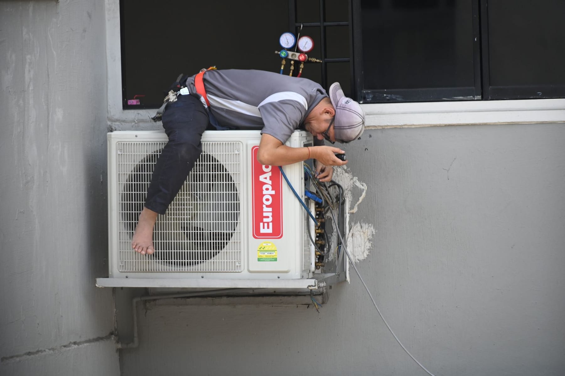 A technician leaning over a wall-mounted condenser, cap down, gauges in hand