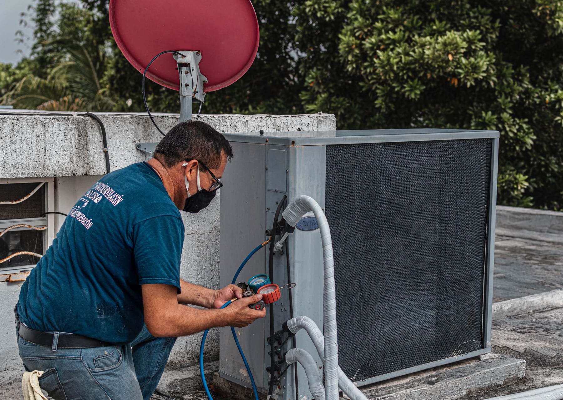 A masked technician in a blue shirt reading gauges on a rooftop condenser