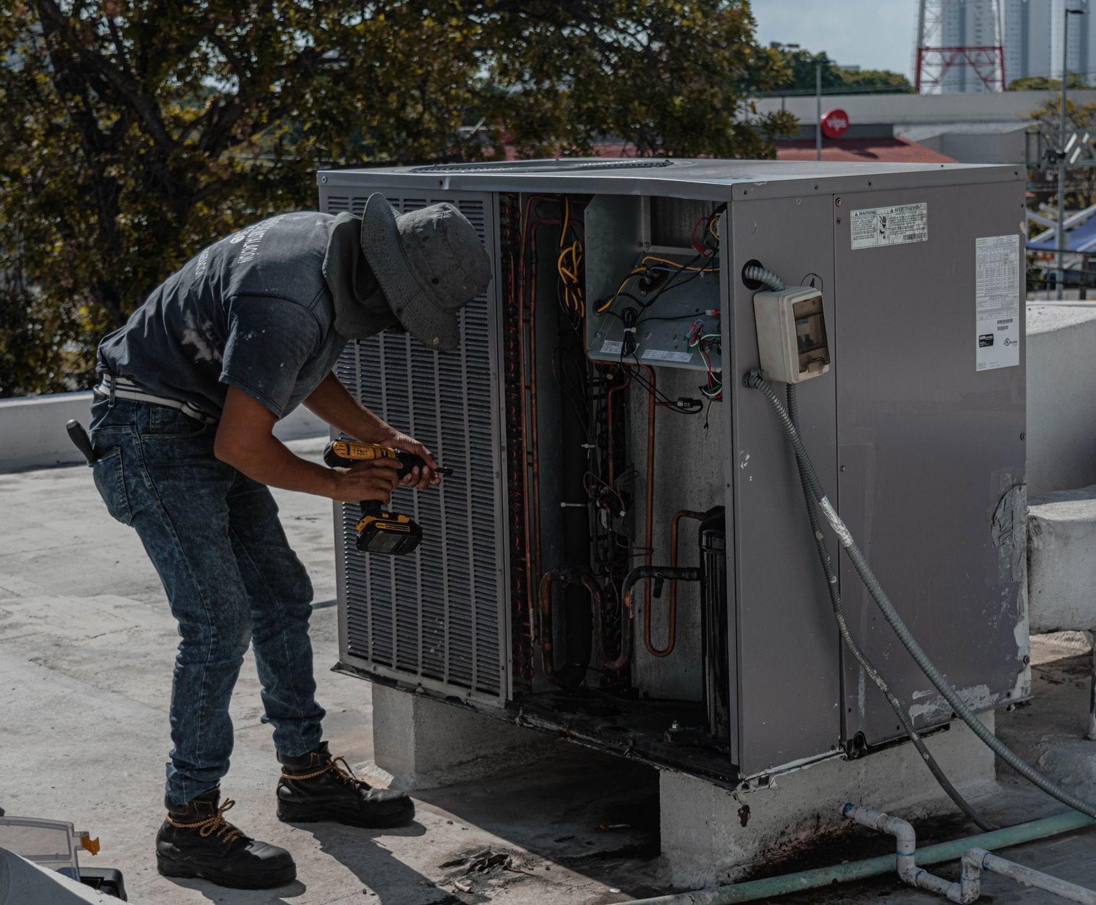 A technician bent into an open rooftop condenser with a drill, sun on his back
